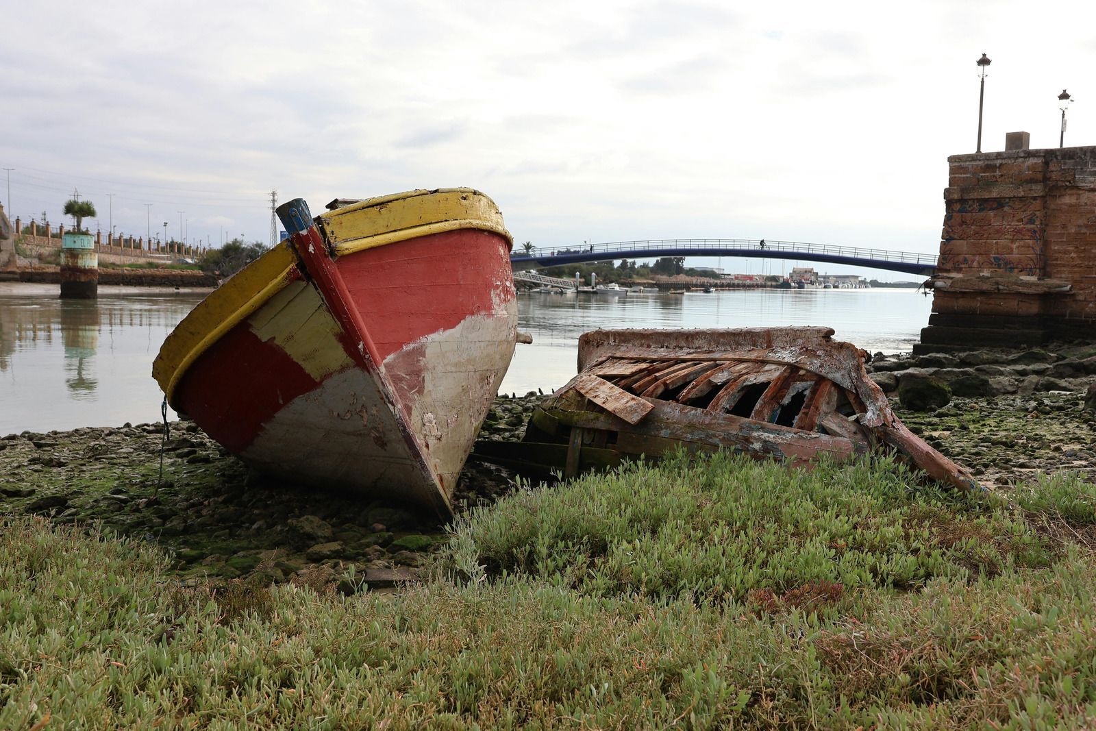 A red-and-yellow boat sits grounded on a muddy riverbank near a bridge and stone wall.