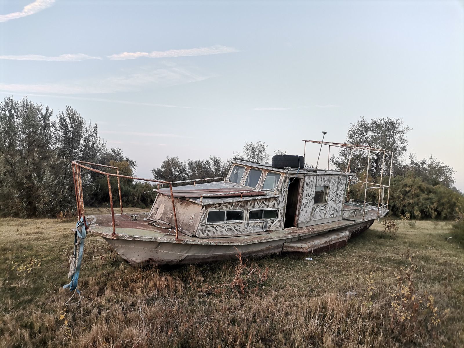 Abandoned white boat in a grassy field, with trees and a pale sky in the background.