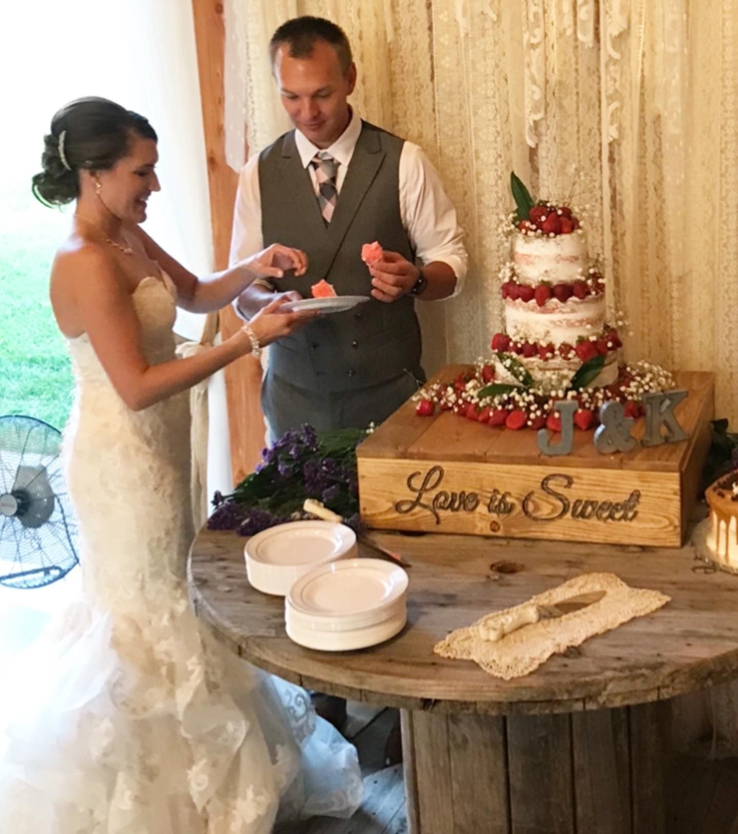 Bride and groom cut wedding cake beside a rustic dessert table with a “Love is Sweet” sign