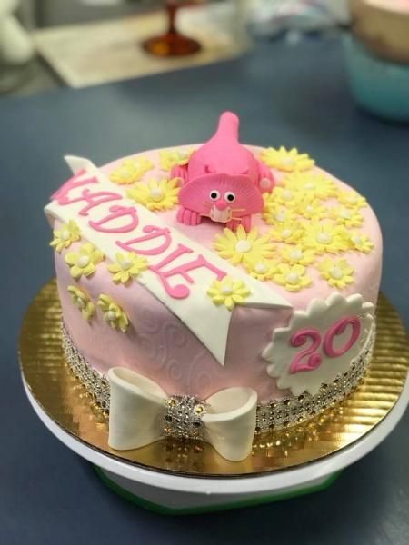 Pink birthday cake with yellow flowers, “Happy” topper, pink character, white bow, and number 20 on a gold board