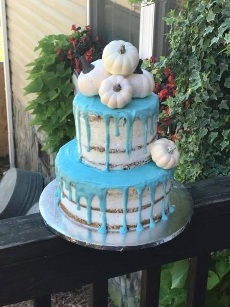 Two-tier blue drip cake on a table, topped with white pumpkins and greenery outdoors