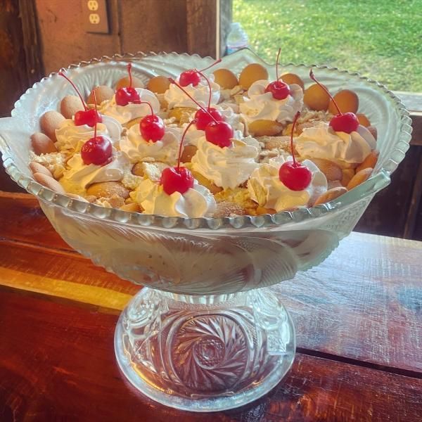 Glass dessert bowl filled with whipped cream, crushed cookies, and cherries on a wooden table by a window