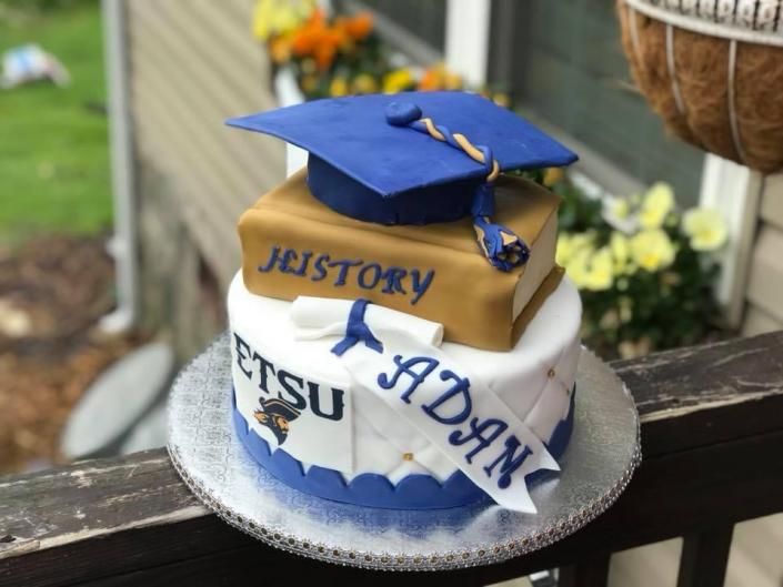 Small graduation-themed cake with blue cap topper and white frosting reading “PSU ADAN” and “HISTORY.”