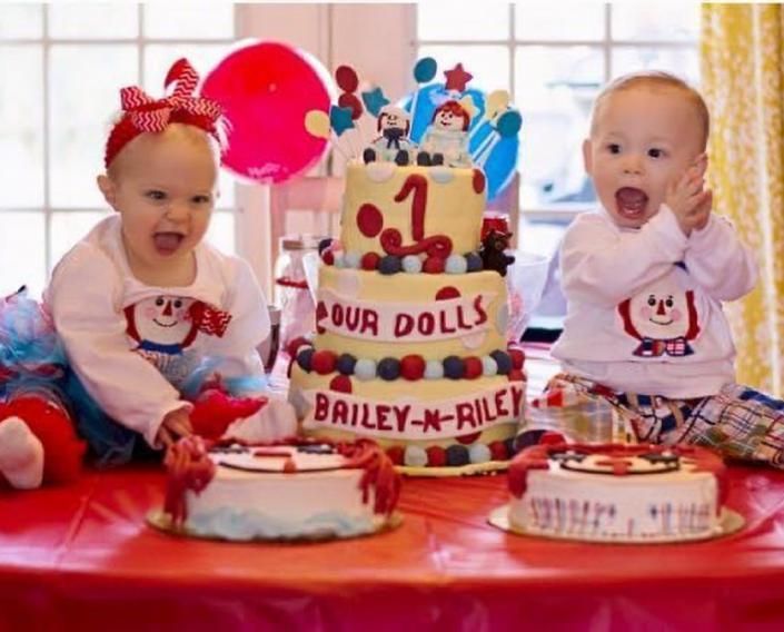 Two babies at a pink birthday cake table with balloons and “OUR DOLL” cake topper, smiling excitedly