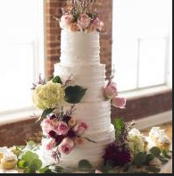 Tall white tiered cake decorated with pink, yellow, and purple flowers on a windowsill