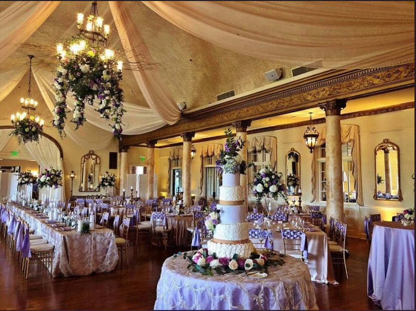 Elegant banquet hall with draped ceiling, chandeliers, floral centerpieces, and round tables set for an event