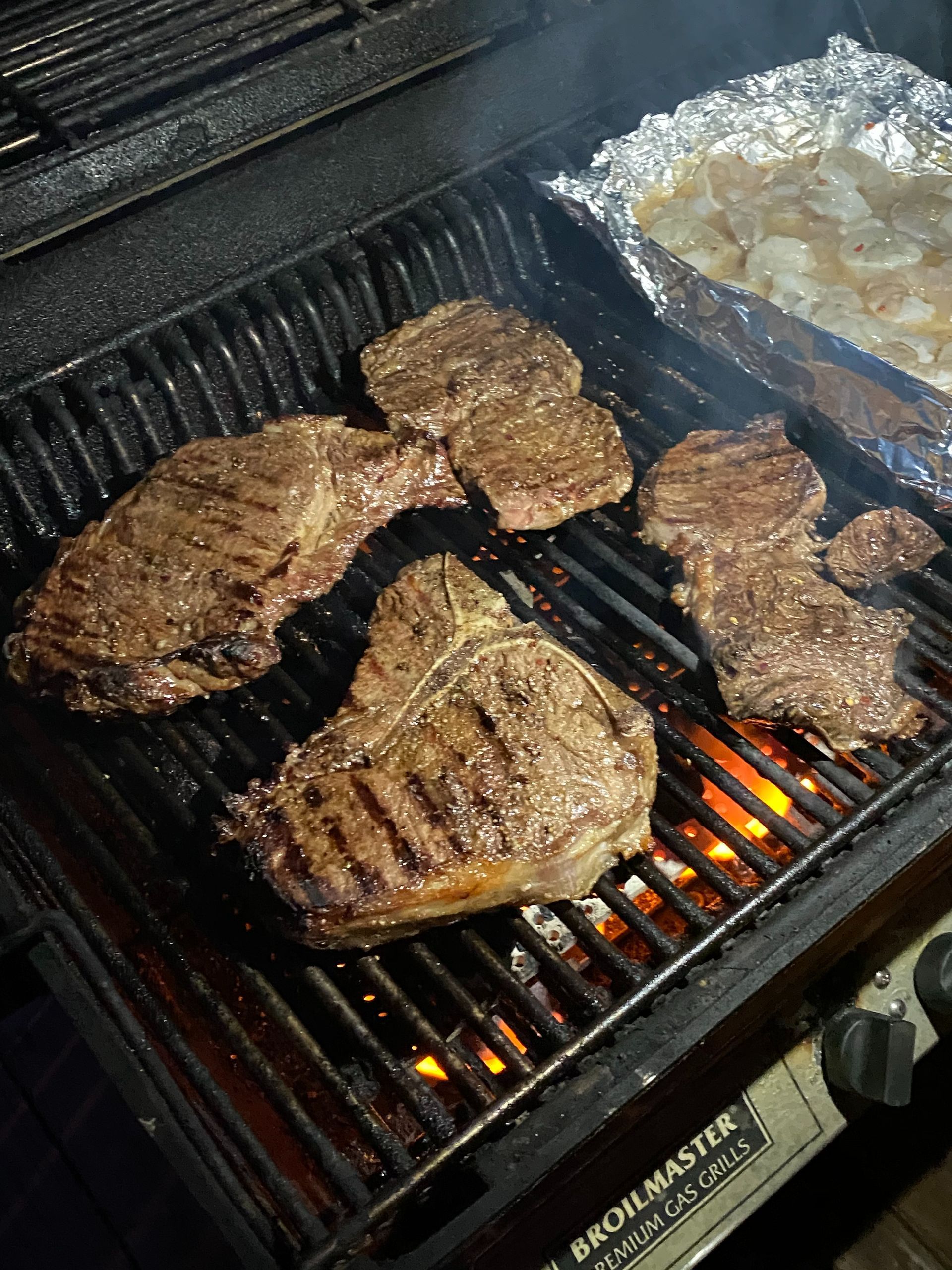Steaks and other meats grilling on a barbecue with visible flames and foil-wrapped food on the side.