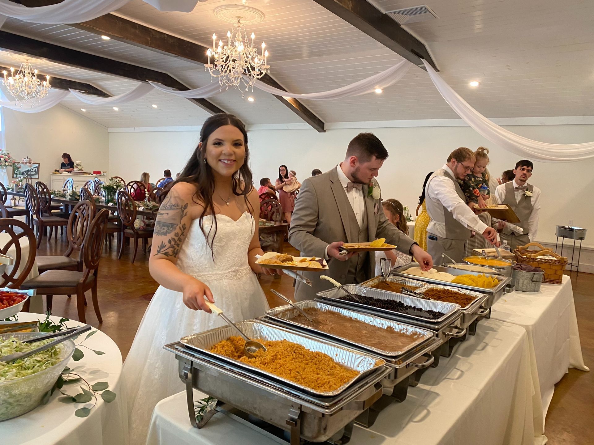 Woman serving food at a buffet in a decorated banquet hall, with trays of hot dishes and guests in the background
