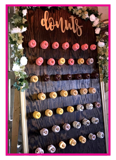 Donut display board with rows of colorful frosted donuts under a “donuts” sign and floral decorations