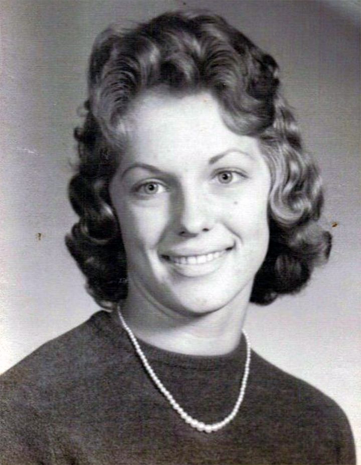 Black-and-white studio portrait of a smiling person with wavy hair and a necklace.