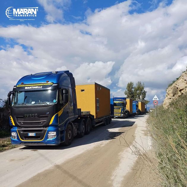 Un camioncino blu di servizio percorre una strada tortuosa vicino a una cittadina collinare e a una montagna rocciosa, con l'oceano in lontananza.