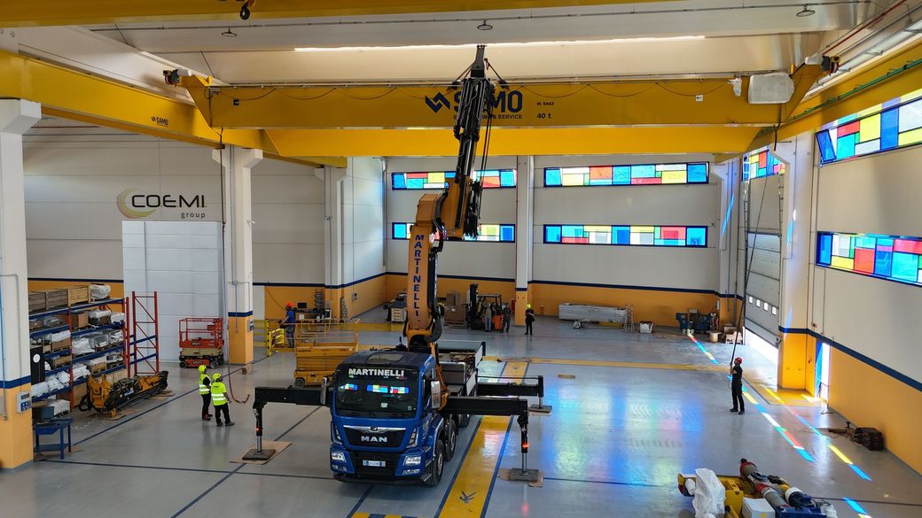 Officina industriale con un camion blu, una gru che solleva carichi in alto e operai all'interno di un grande magazzino.