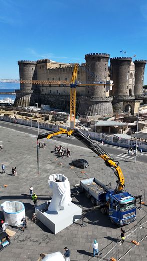 Fortezza in pietra con torri accanto a una piazza, una gru gialla e una grande scultura bianca in primo piano