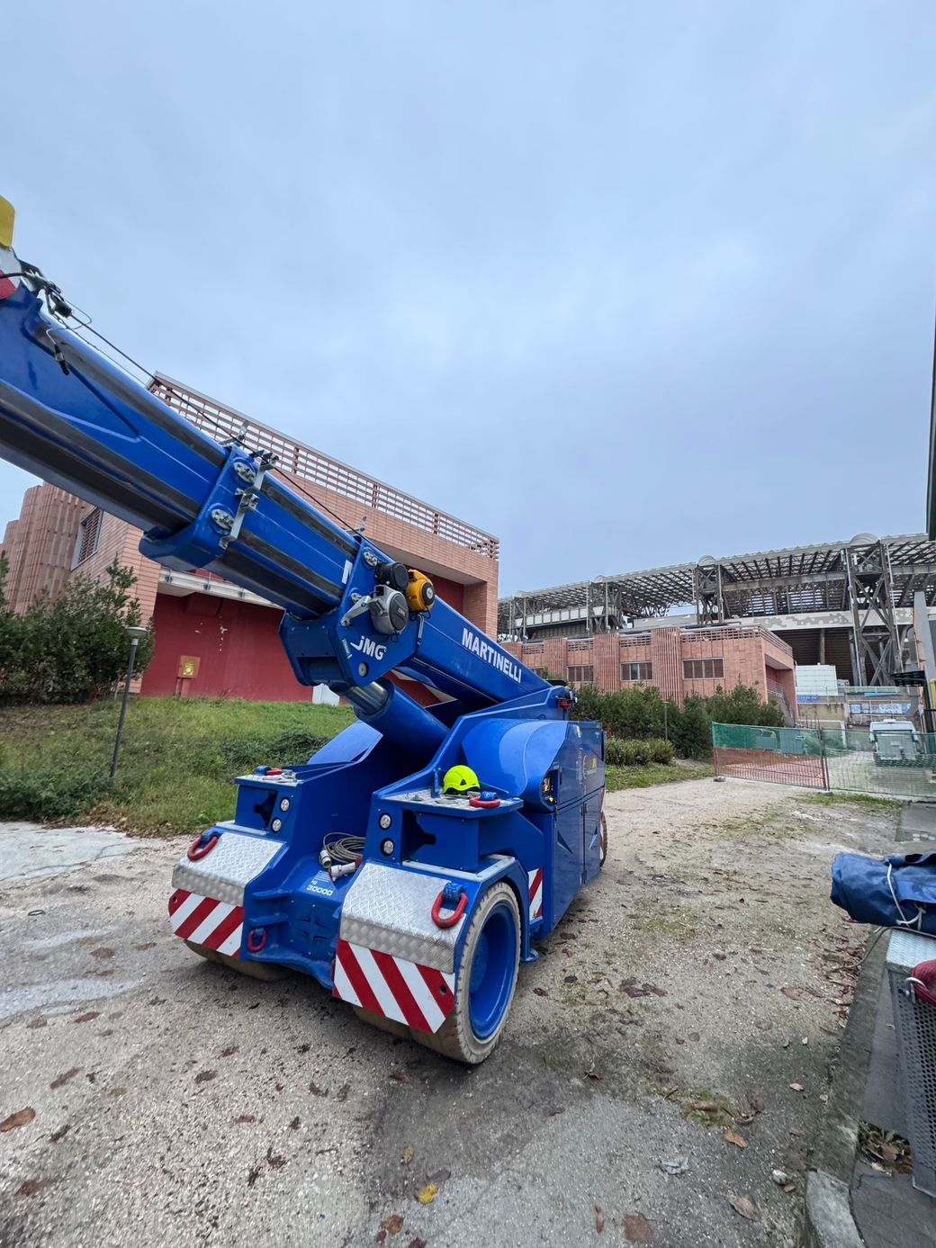 Una gru blu è parcheggiata su un piazzale ghiaioso accanto a un edificio danneggiato, con il braccio esteso verso l'alto.