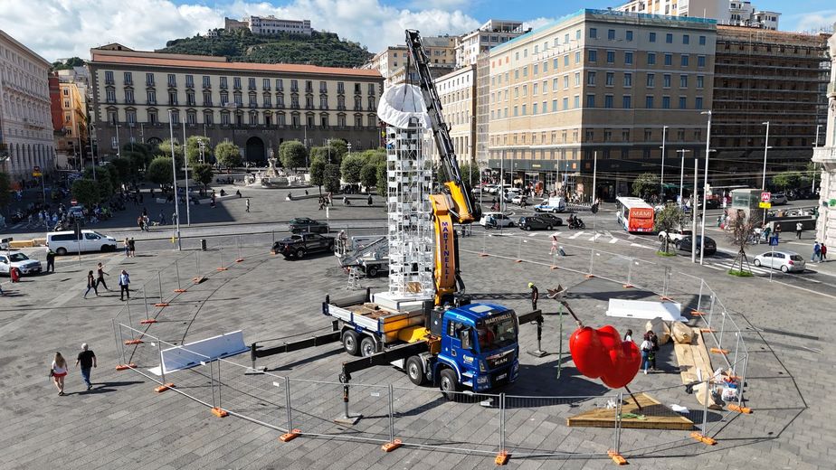 Ampia piazza cittadina con persone sparse, attrezzature da cantiere e una grande scultura rossa vicino agli edifici circostanti.