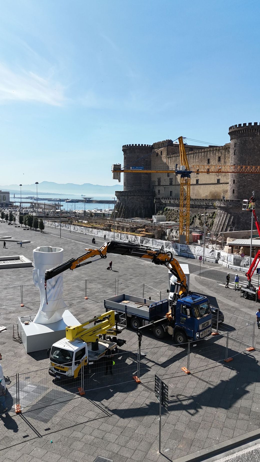 Una gru gialla accanto a un muro di pietra del porto, con persone e attrezzature su un lungomare soleggiato.