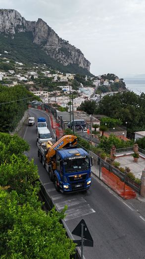 Un camioncino blu di servizio percorre una strada tortuosa vicino a una cittadina collinare e a una montagna rocciosa, con l'oceano in lontananza.