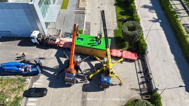 Una gru da cantiere in una piazza sul lungomare, accanto a un edificio storico in pietra, sotto un cielo azzurro e limpido.
