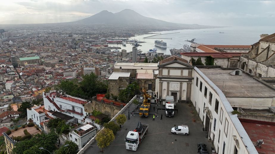 Veduta panoramica di una città costiera con montagne, una baia e una strada collinare fiancheggiata da edifici e automobili.