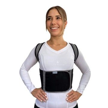 Mujer con un corsé ortopédico negro sobre una camisa blanca, vista de espaldas sobre un fondo blanco.