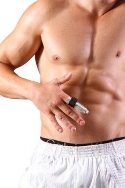 Persona sin camisa sosteniendo una jeringa en su abdomen, vistiendo pantalones cortos blancos sobre un fondo liso.