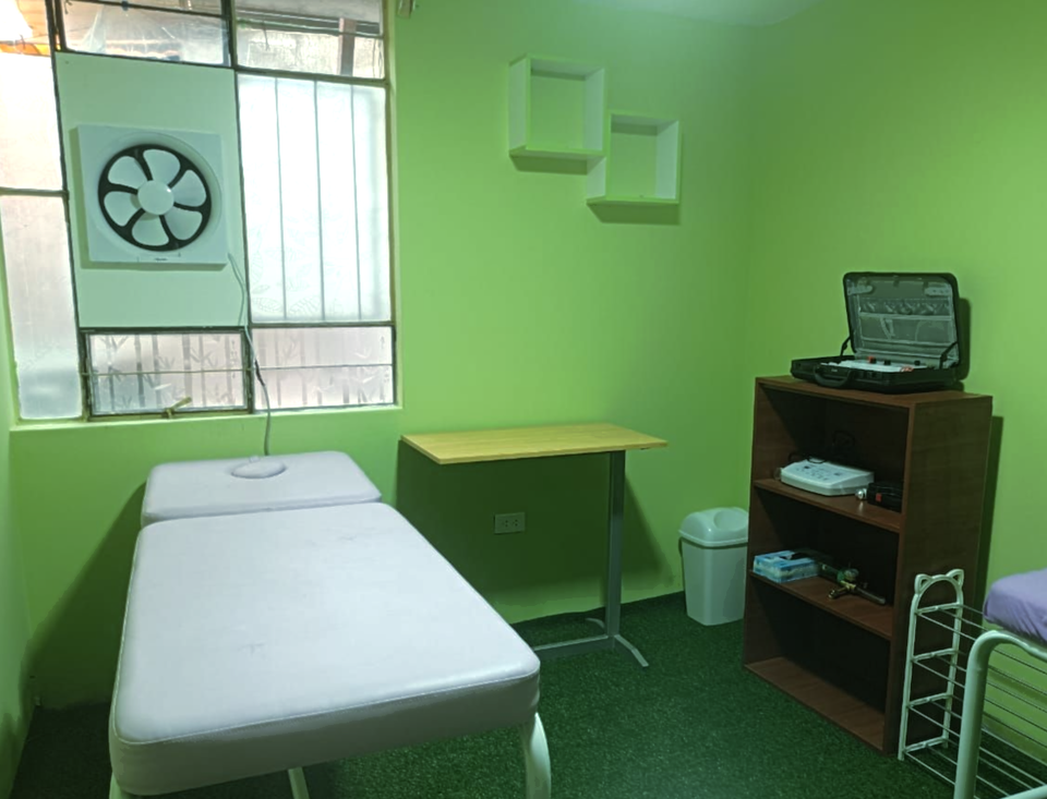 Sala de examen verde con camilla de tratamiento, escritorio, ventilador de pared y luz de ventana.