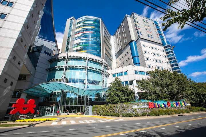 Modern multi-story hospital building with glass entrance and red signage under a blue sky