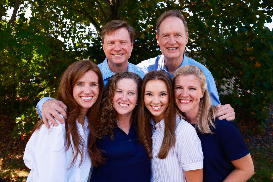 Family of six posing outdoors in front of greenery, smiling and standing close together.
