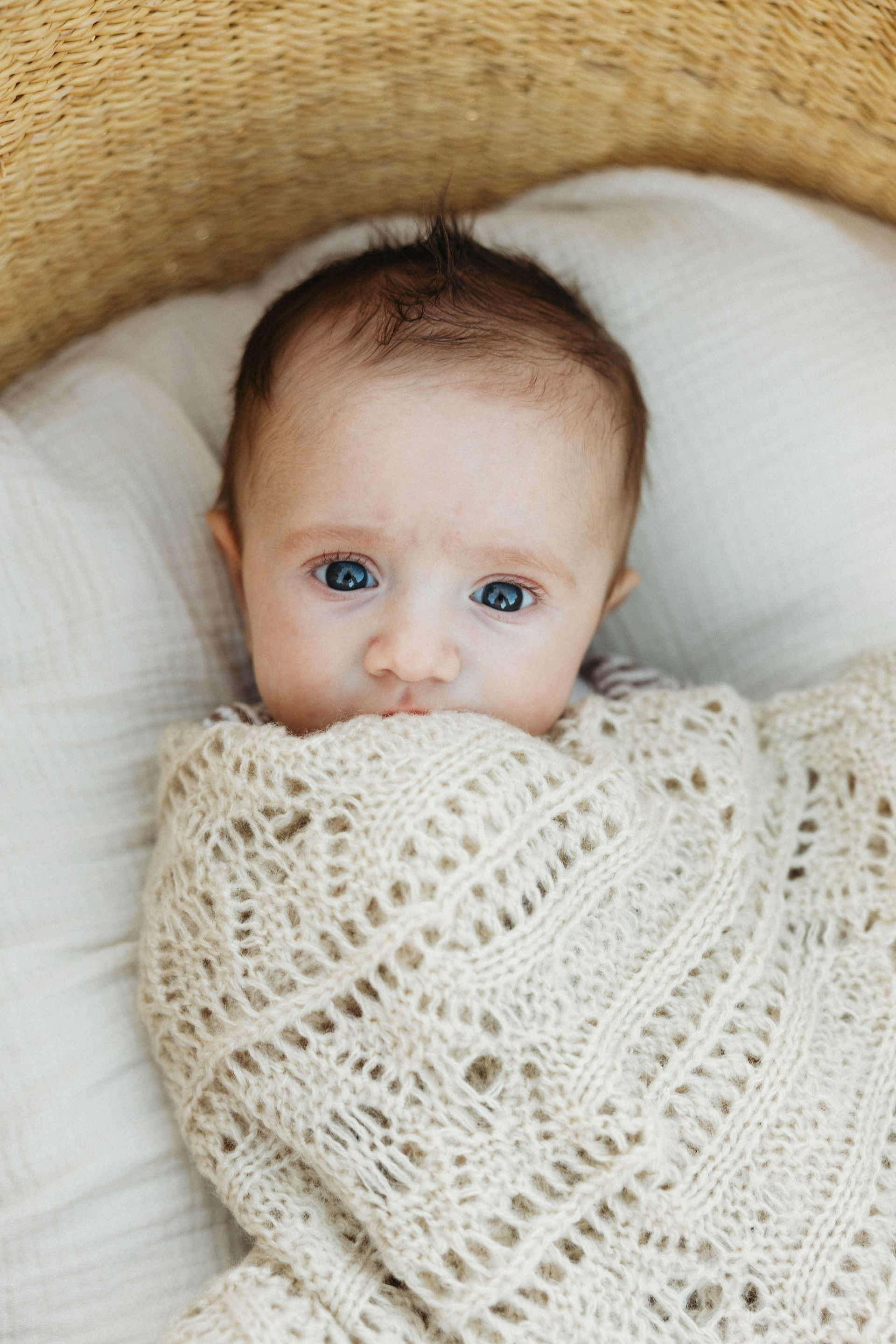 Baby wrapped in a cream knit blanket, lying in a white basket with a woven tan backdrop.