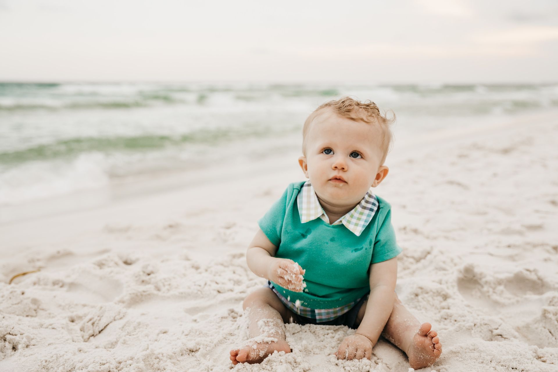 Toddler sitting on a sandy beach in a teal shirt, with ocean waves in the background.