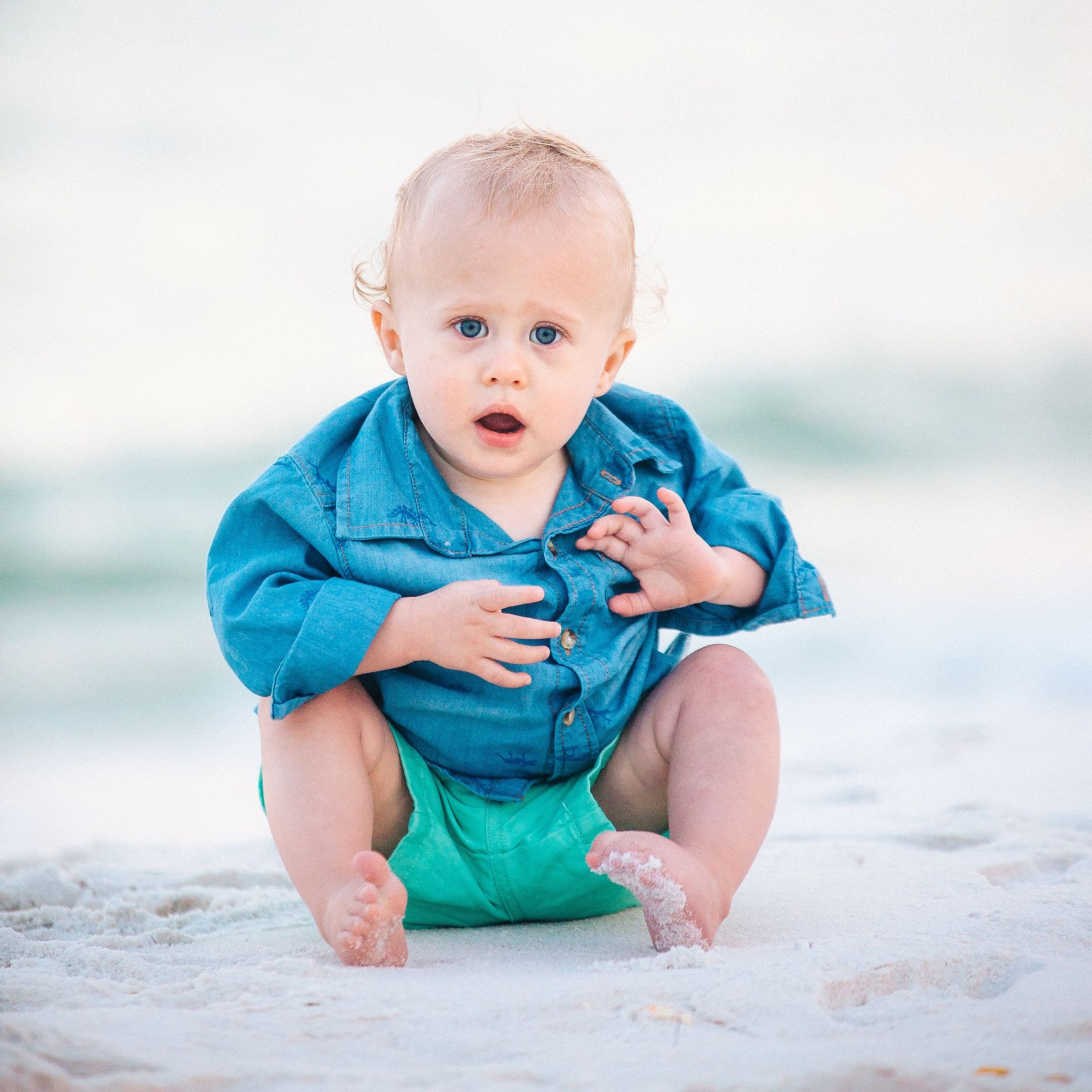 Baby sitting on a beach in a blue shirt and green shorts, looking at the camera.