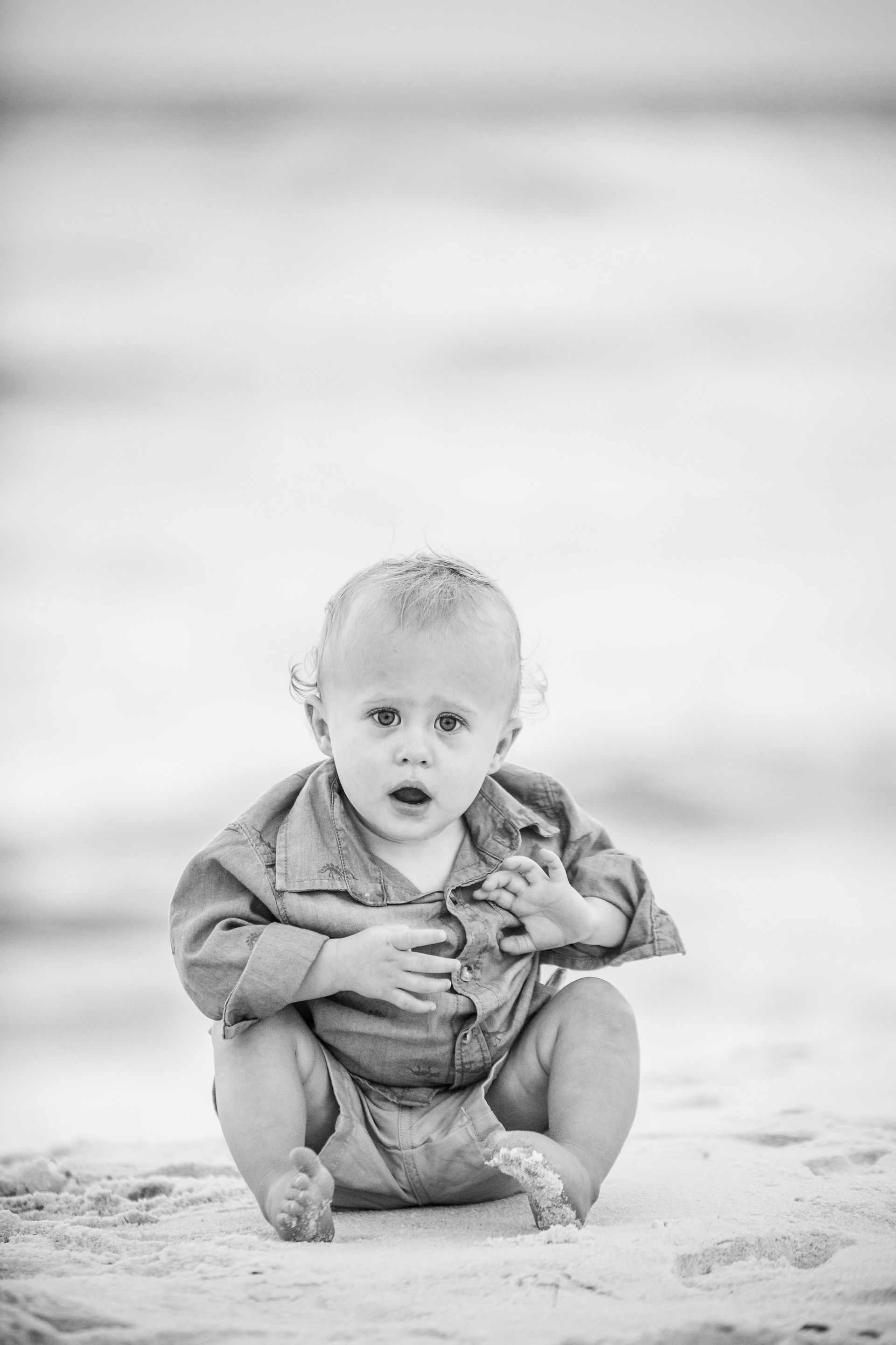 A baby sitting on a sandy beach, holding a stuffed toy and looking toward the camera.