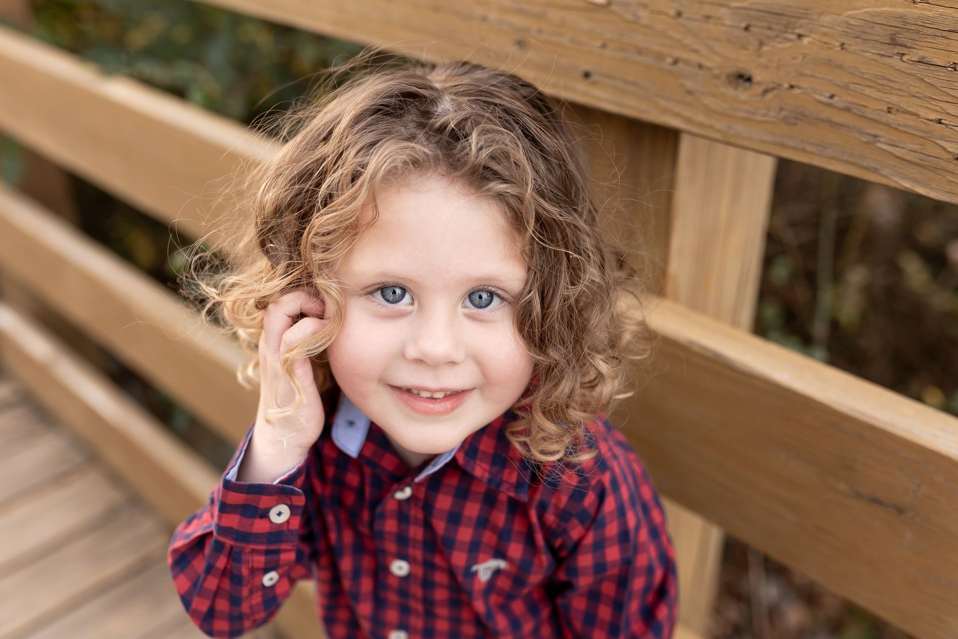 Child with curly hair in a red plaid shirt leaning by a wooden fence and smiling at the camera
