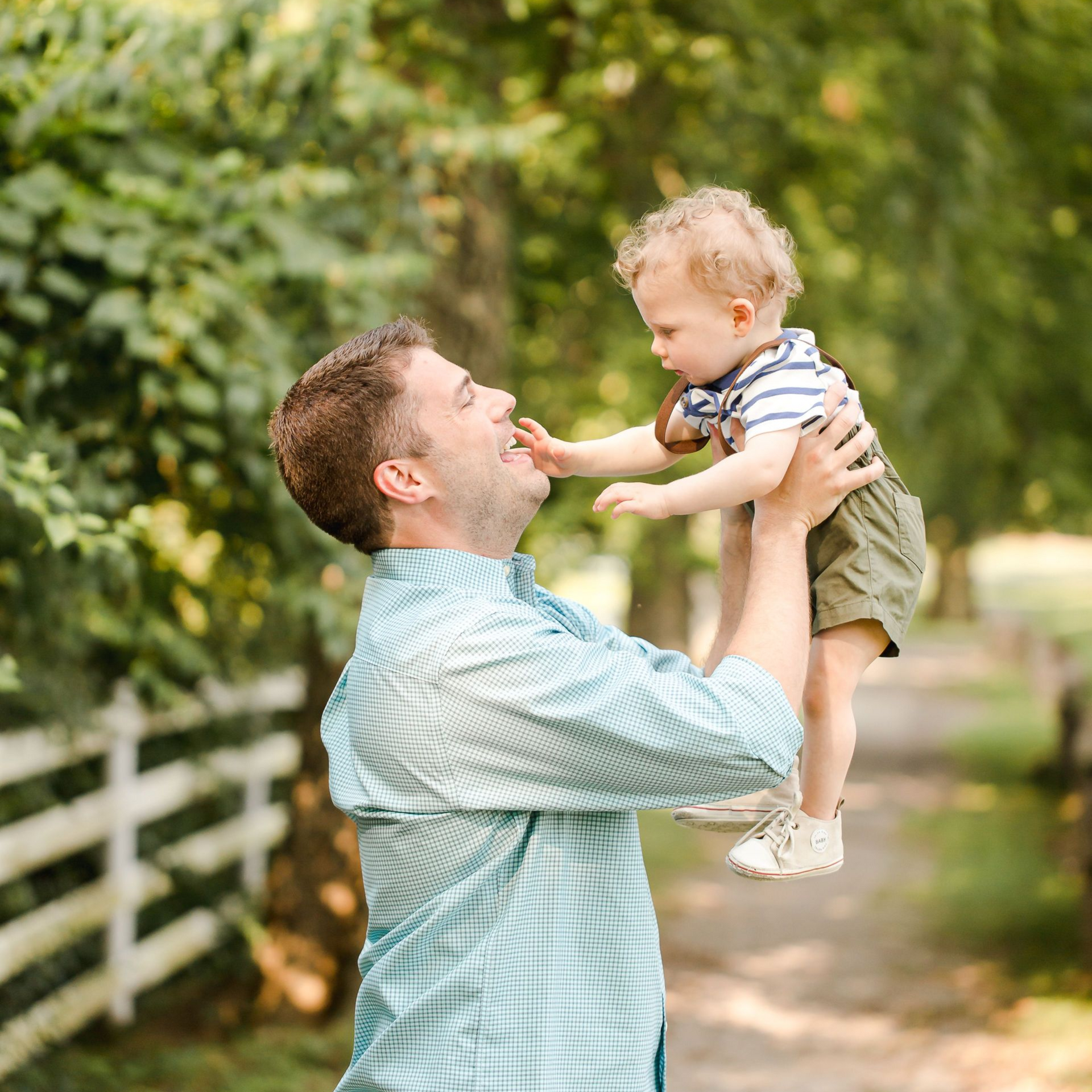 Man lifting a smiling toddler outdoors on a sunlit path with greenery and a white fence nearby