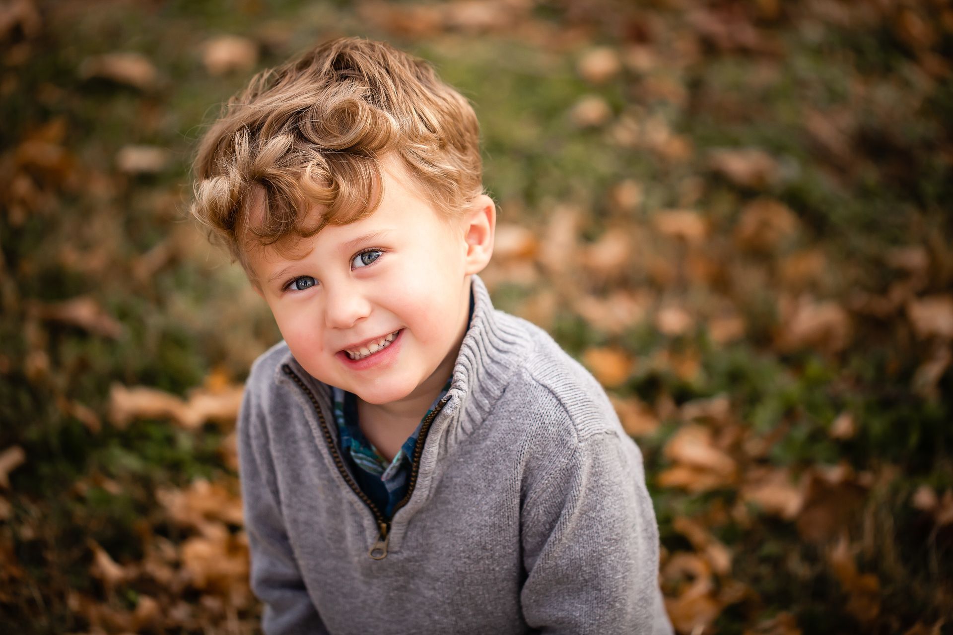 Smiling child in a gray sweater outdoors with blurred autumn leaves in the background