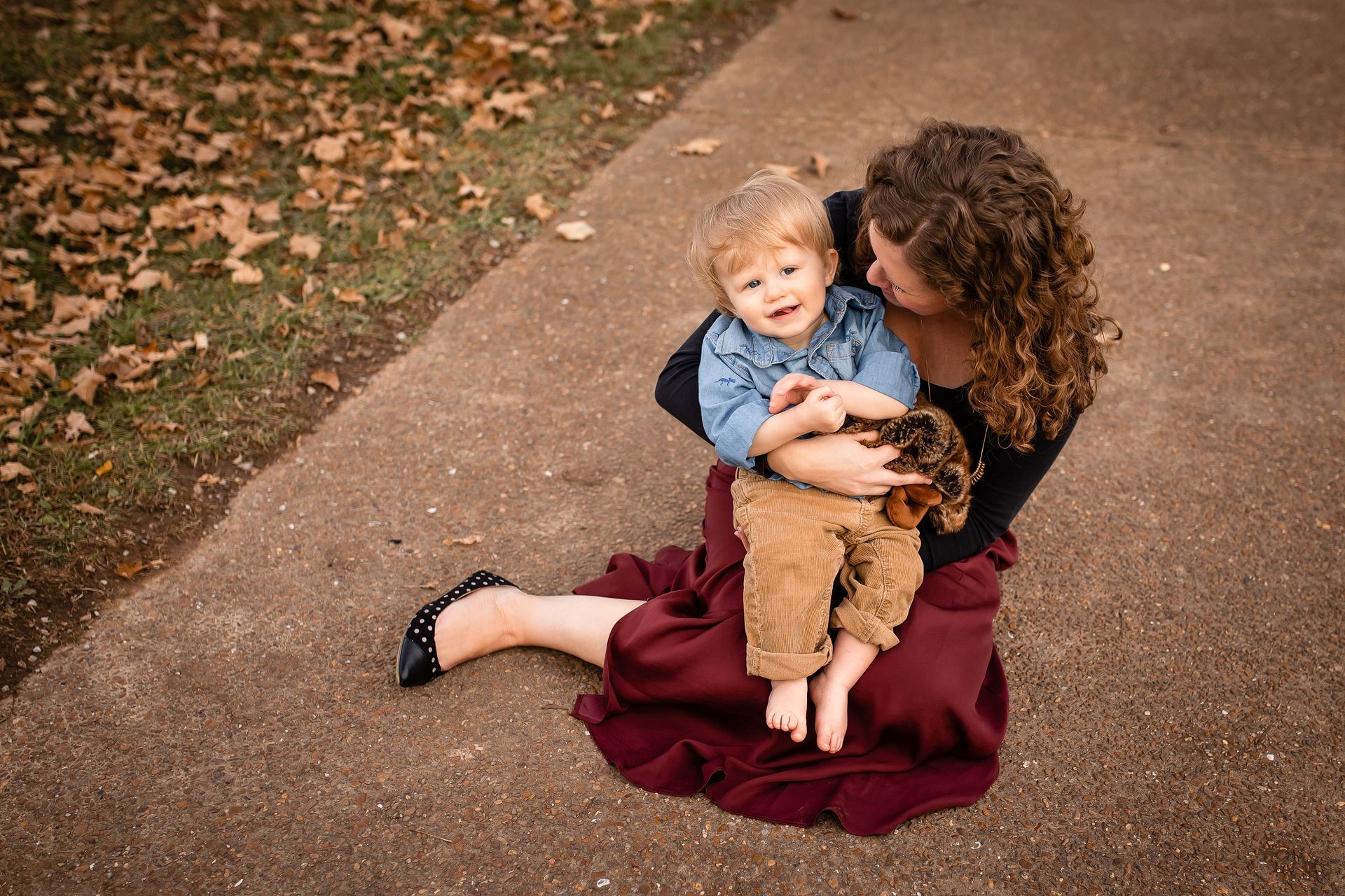 Woman in burgundy dress holding a toddler on a paved path with fallen leaves nearby