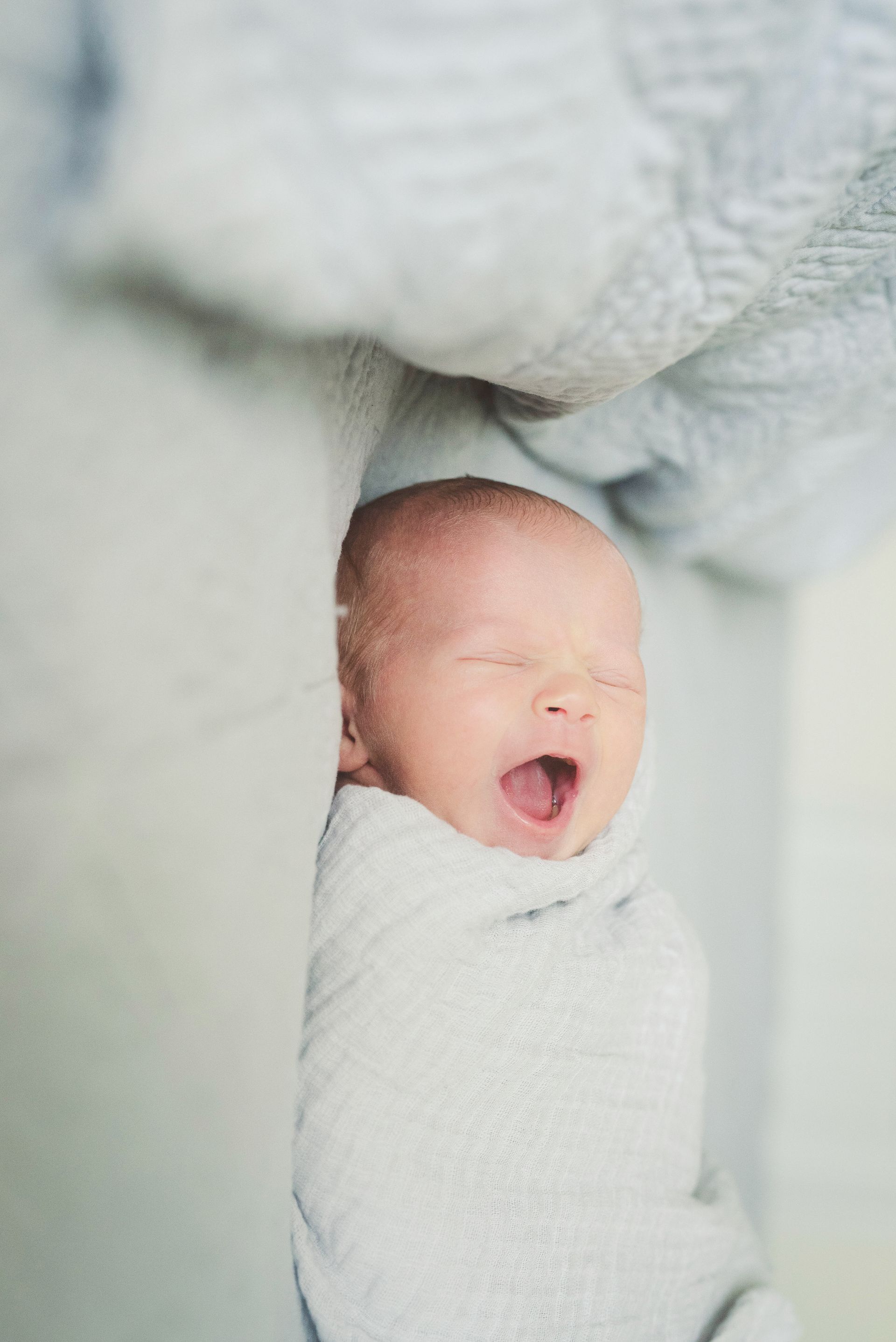 Newborn wrapped in a white blanket, yawning with eyes closed.