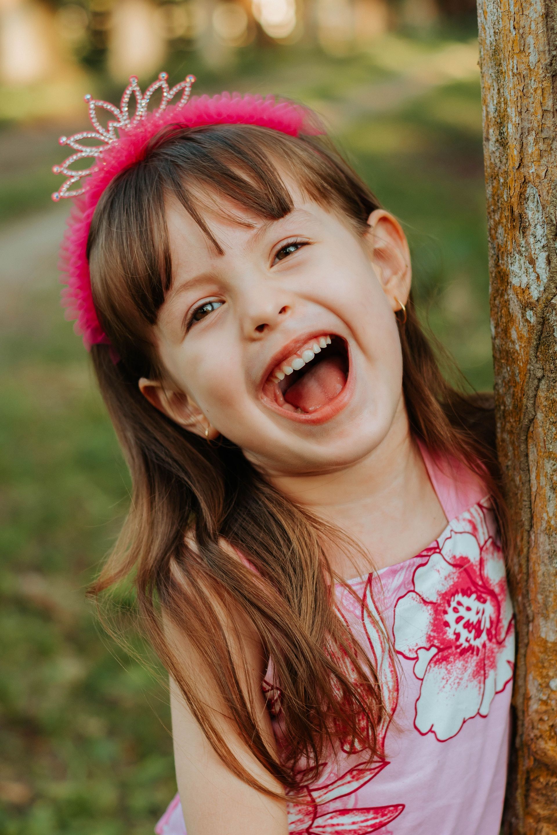 Smiling child in a pink dress and headband, laughing outdoors beside a tree