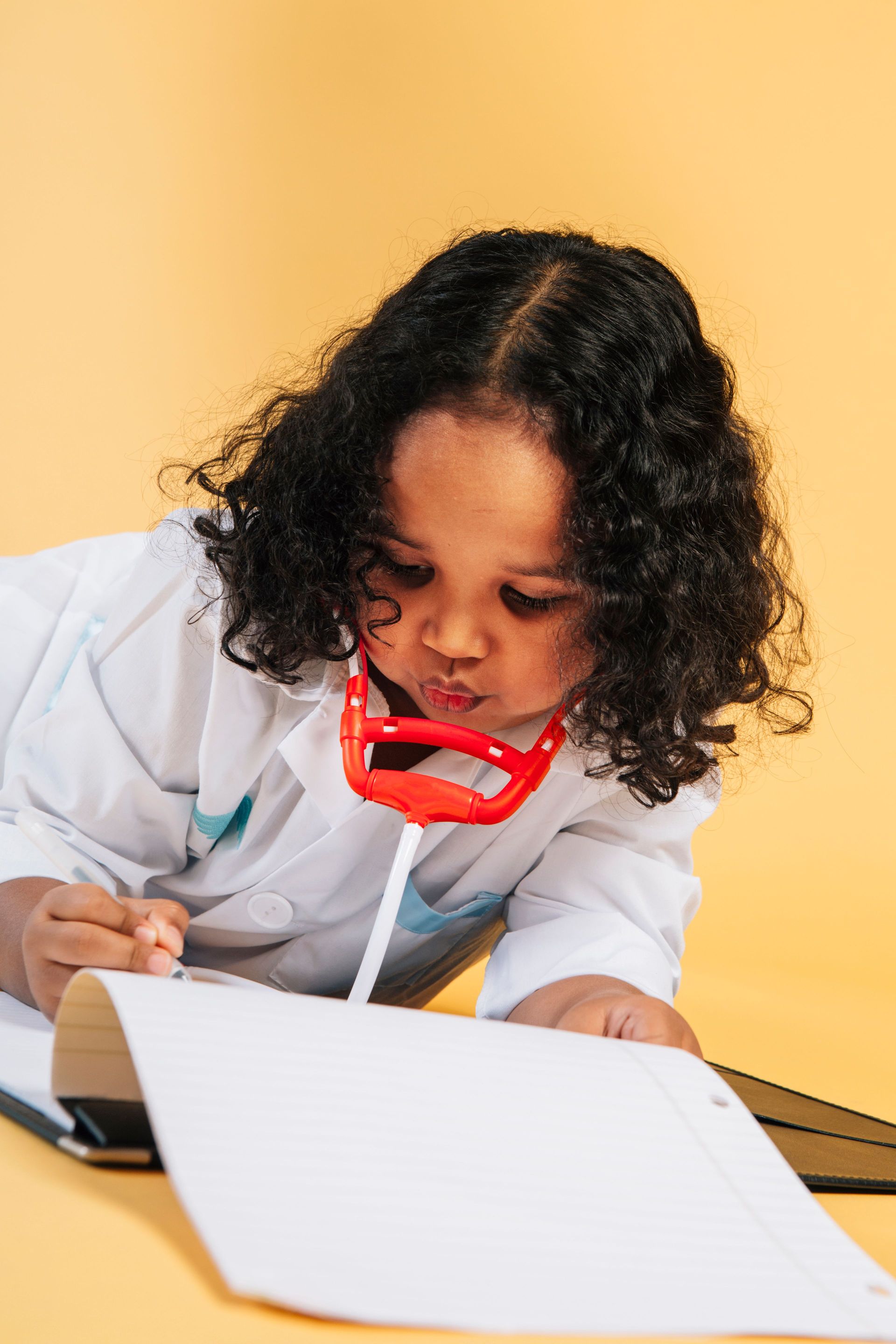 Child writing with red glasses on a white notebook against a yellow background