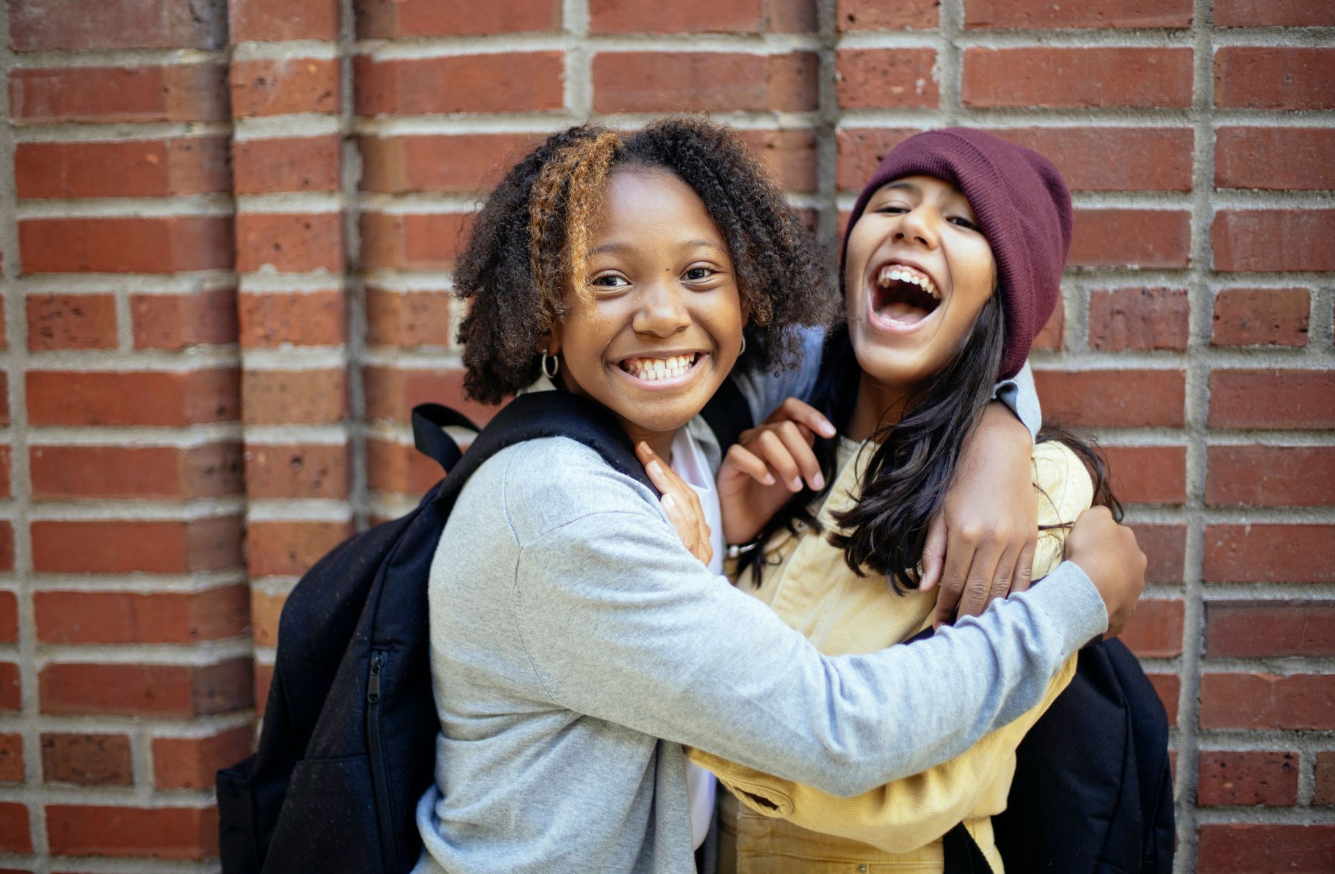 Two friends hugging and laughing in front of a brick wall, both wearing backpacks.