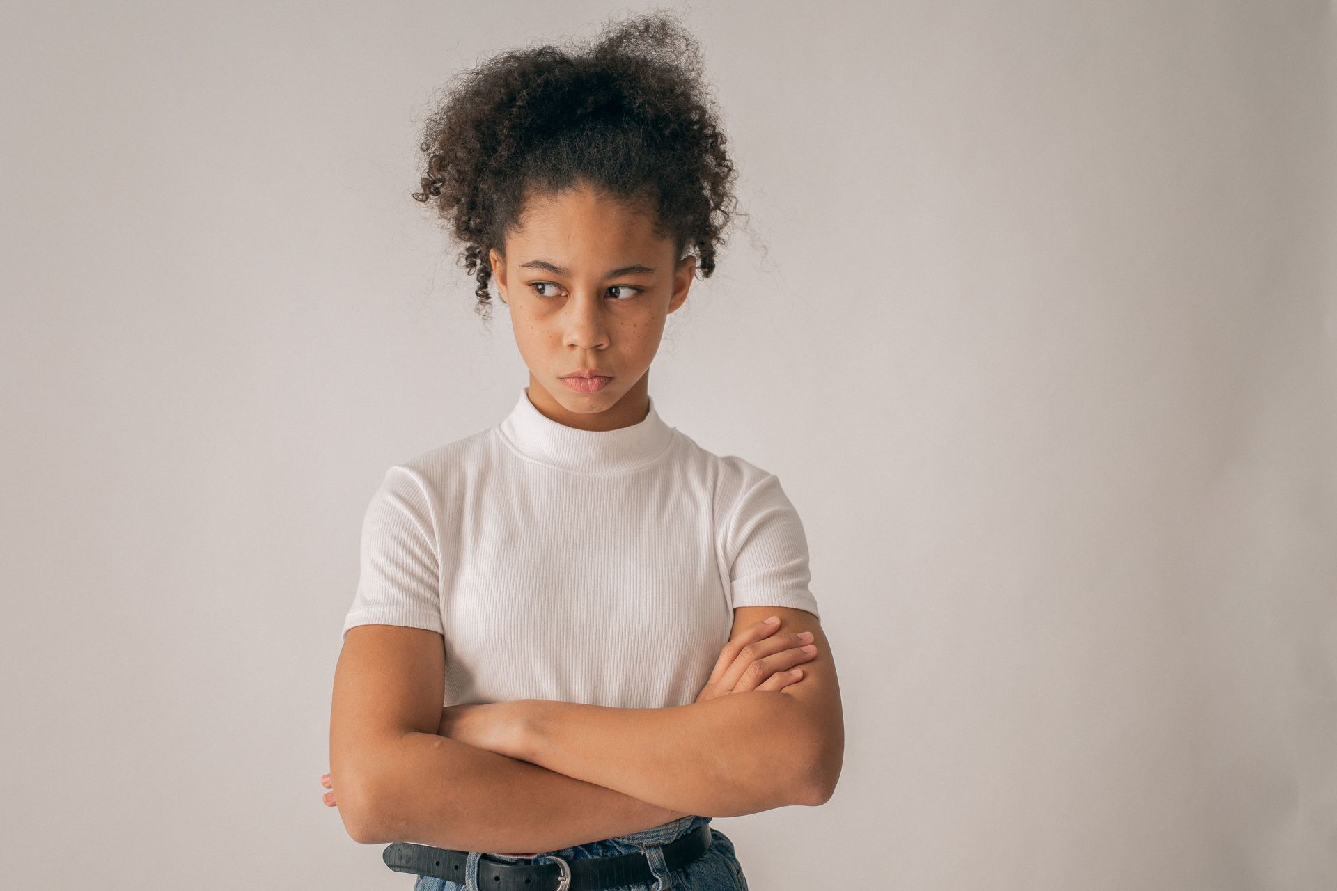 Woman with curly hair standing against a plain wall, arms crossed, wearing a white ribbed shirt and jeans.