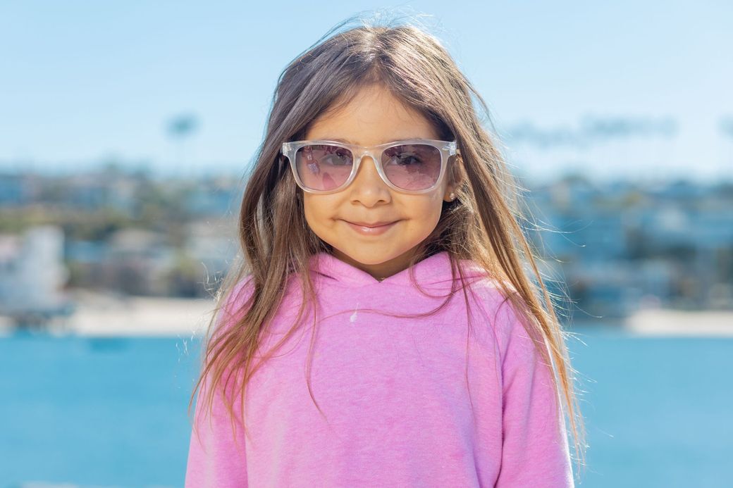 Girl in pink sweater and sunglasses standing outdoors with a blurred waterfront background