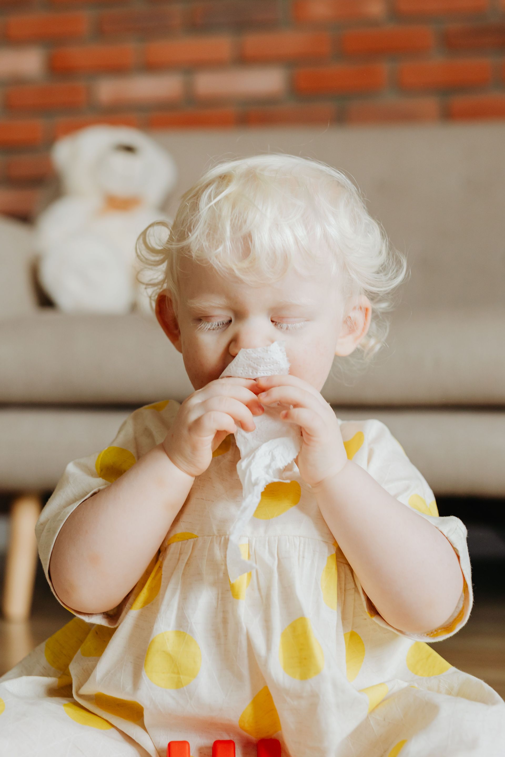 Toddler in a yellow-and-white dress holding a snack indoors, sitting on a couch with a stuffed toy in the background