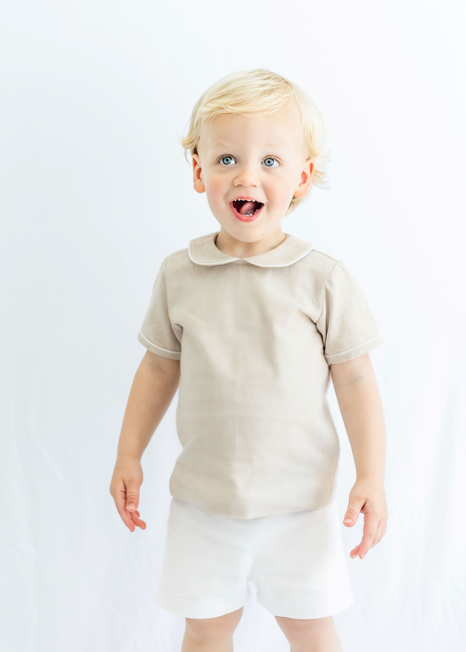 Young child in a beige shirt and white shorts standing against a white background, smiling.