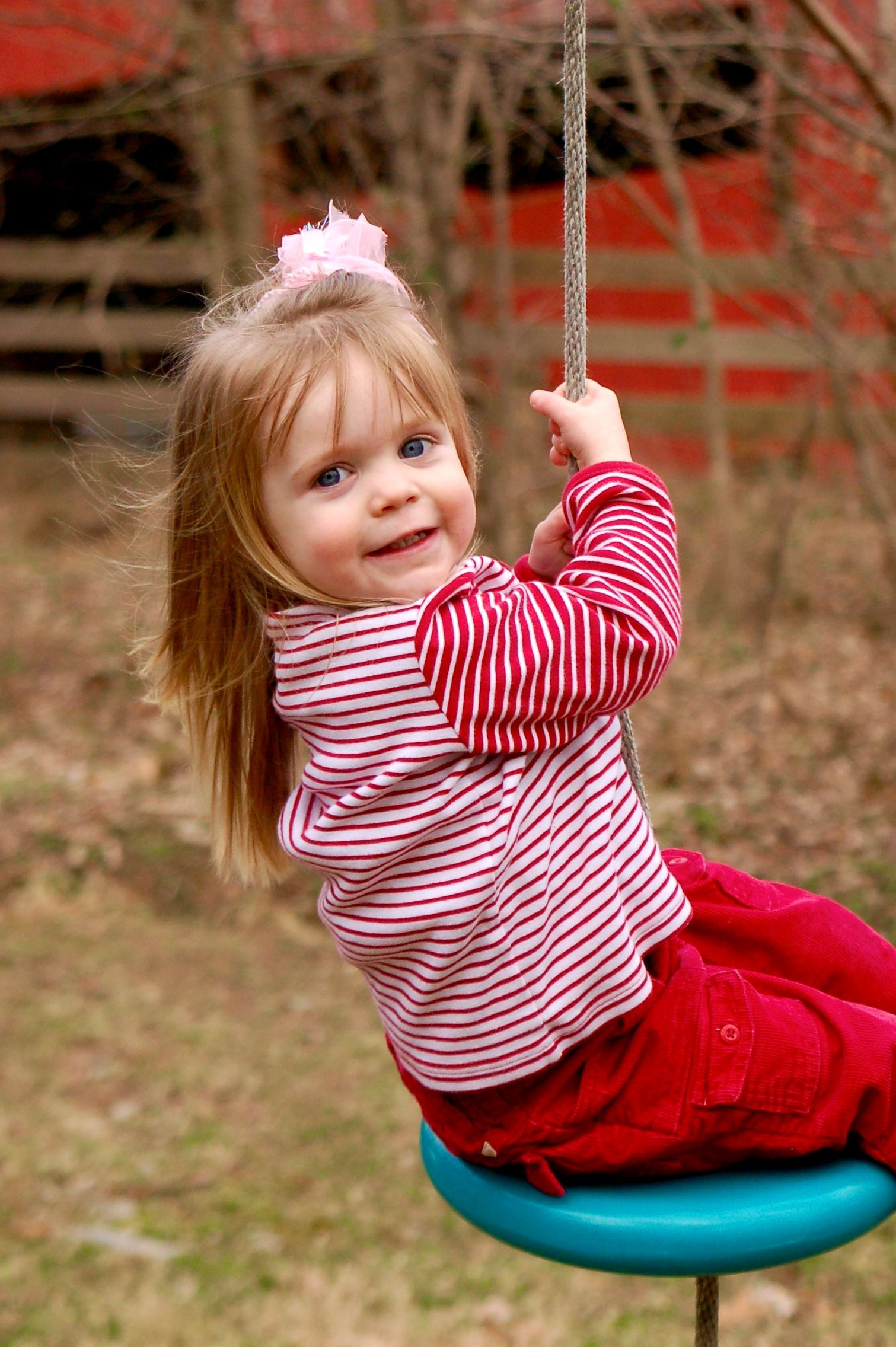 Child smiling on a swing outdoors, wearing a red striped shirt and bright red pants.