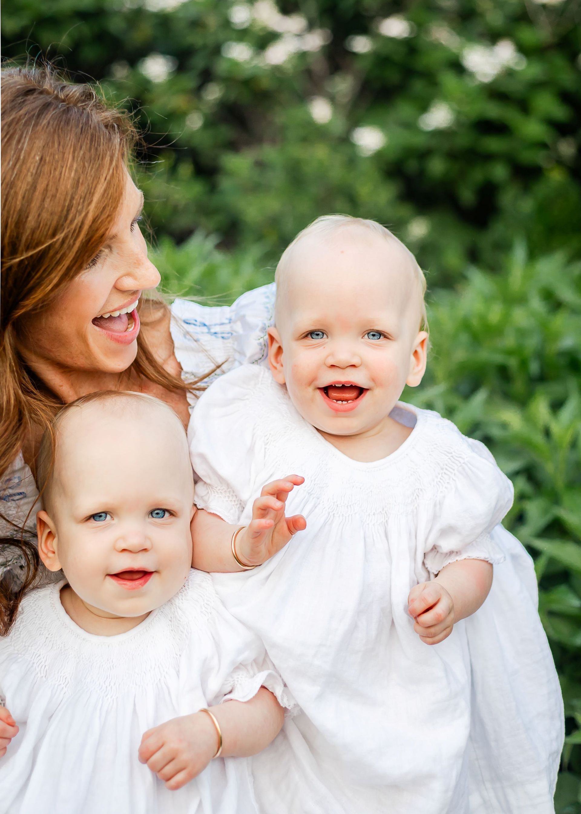 Woman with two smiling babies in white outfits outdoors, with green foliage in the background