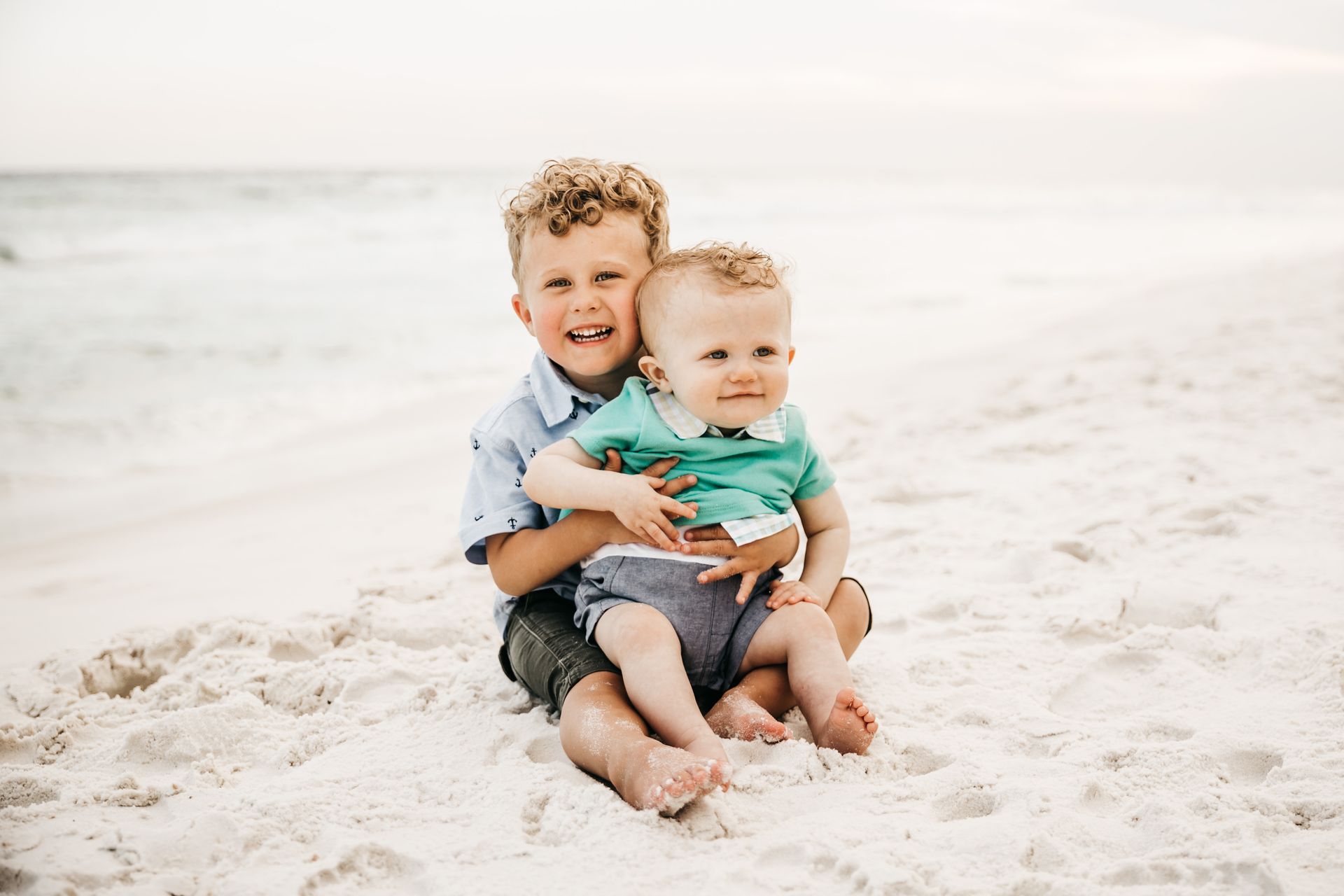 Two small children sitting together on a bright sandy beach, smiling and hugging