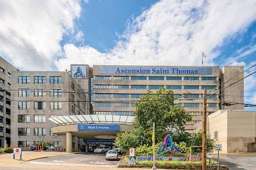 Entrance of Ascension Saint Thomas hospital with tall glass facade, trees, and parked cars under a blue sky