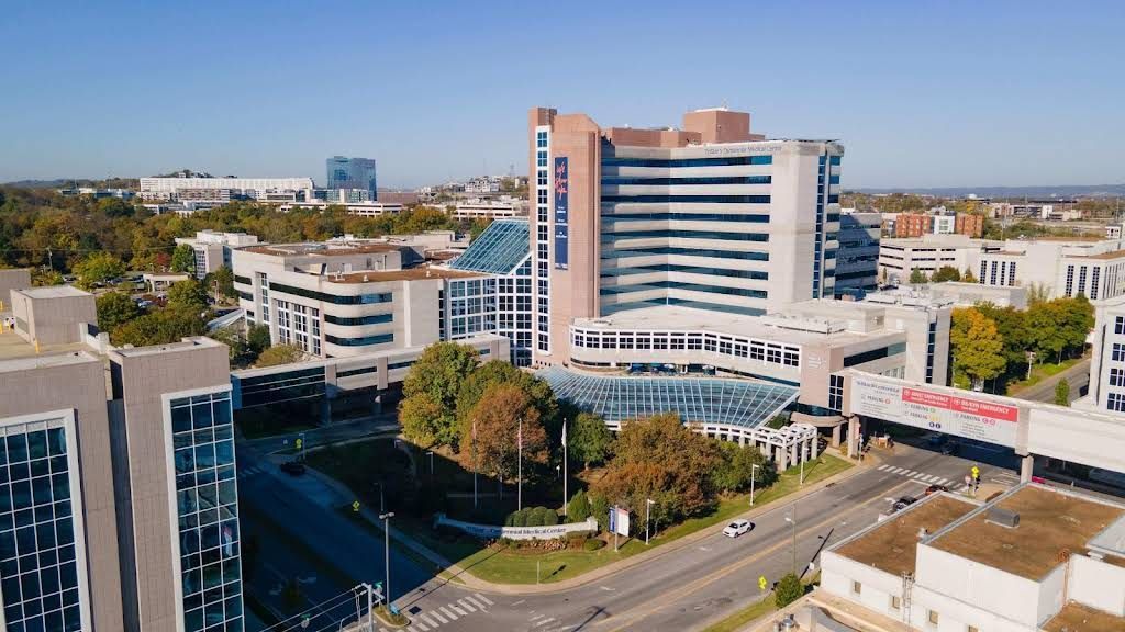 Aerial view of a modern hospital campus with white and pink buildings, trees, and surrounding roads.
