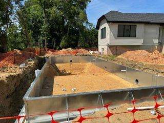 Concrete pool foundation being built beside a house, with dirt excavation and orange safety fencing.